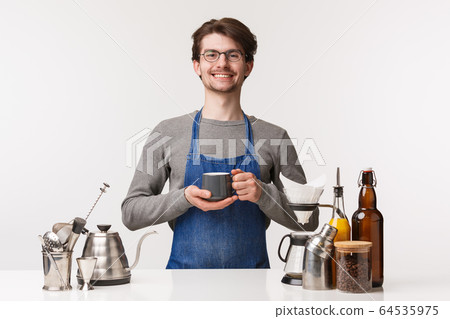 Barista, cafe worker and bartender concept. Portrait of cheerful pleasant young male employee in apron invite everyone have delicious drink, making coffee, holding tea and smiling camera Barista, cafe worker and bartender concept. Portrait of cheerful pleasant young male employee in apron invite everyone have delicious drink, making coffee, holding tea and smiling camera 64535975