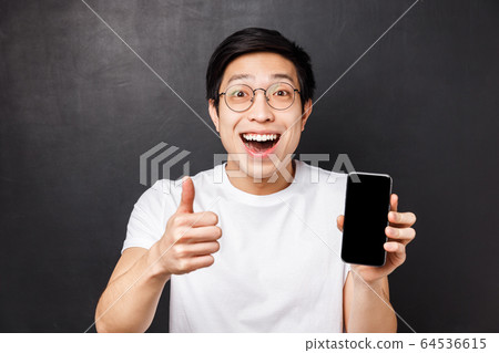 Technology, messaging and people concept. Close-up portrait of excited, happy young asian man show thumb-up and mobile phone display, smiling astonished, rate good app, recommend subscription Technology, messaging and people concept. Close-up portrait of excited, happy young asian man show thumb-up and mobile phone display, smiling astonished, rate good app, recommend subscription 64536615