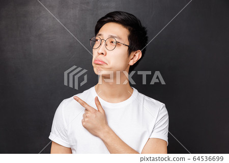 Close-up portrait of jealous and gloomy asian young man feel regret, look at something upsetting, pouting pointing finger left, desire have something but cant afford it, black background 64536699