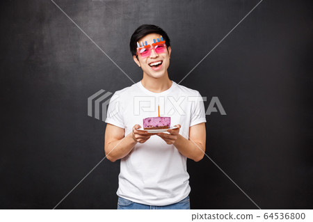 Birthday, celebration and party concept. Enthusiastic cute asian guy celebrating b-day, tilt head and look happy camera with pleased smile, hold cake on plate with lit candle, making wish 64536800