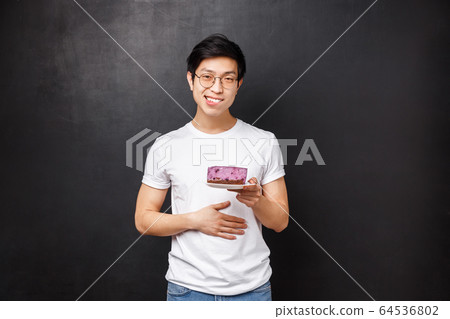 Portrait of pleased and staffed young cute asian man with piece of cake on plate, rubbing his belly and smiling satisfied, ate tasty dessert, delighted after having delicious meal, black background Portrait of pleased and staffed young cute asian man with piece of cake on plate, rubbing his belly and smiling satisfied, ate tasty dessert, delighted after having delicious meal, black background 64536802