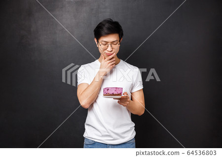 Portrait of thoughtful and doubtful asian guy looking hesitant at piece of cake, thinking eat or leave it in the fridge, standing over black background, being on diet, allergic on ingridients 64536803