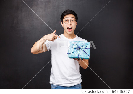 Celebration, holidays and lifestyle concept. Portrait of excited and curious cute asian guy asking whats inside gift box, celebrating b-day party holding present and pointing finger at it 64536807