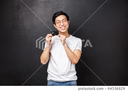 Bank, finance and payment concept. Portrait of silly and happy asian man in t-shirt, close eyes satisfied introducing his first credit card, ready go shopping and waste some money, black background 64536829