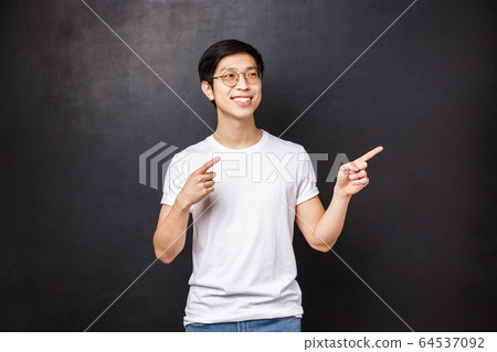 Handsome stylish young asian guy in white shirt, sunglasses pointing looking right side copy space, pleased smile, found exactly what he wanted, choosing this product, black background 64537092