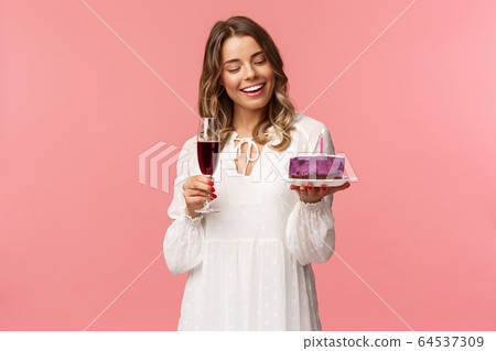 Holidays, spring and party concept. Portrait of tender, elegant young blond woman holding glass of wine and cake with lit candle, smiling pleased, celebrating birthday, pink background Holidays, spring and party concept. Portrait of tender, elegant young blond woman holding glass of wine and cake with lit candle, smiling pleased, celebrating birthday, pink background 64537309