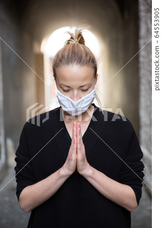 Coronavirus outbreak. Young caucasian woman wearing medical protection face mask praying over coronavirus global pandemic, for salvation of humanity, health, anxiety and depression reduction Coronavirus outbreak. Young caucasian woman wearing medical protection face mask praying over coronavirus global pandemic, for salvation of humanity, health, anxiety and depression reduction 64553905