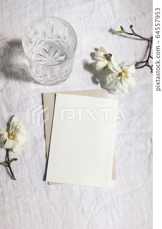 Wedding stationery set. Mock-up scene with blank paper greeting cards, envelope and glass of water on linen tablecloth background. White magnolia stellata tree branches in sunlight.Vertical still life Wedding stationery set. Mock-up scene with blank paper greeting cards, envelope and glass of water on linen tablecloth background. White magnolia stellata tree branches in sunlight.Vertical still life 64557953