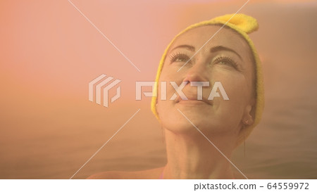 Close up caucasian woman in a swim in hot saline mineral water bath at a traditional spa outdoor with soft magic pink lighting steam. Geothermal spa. 64559972