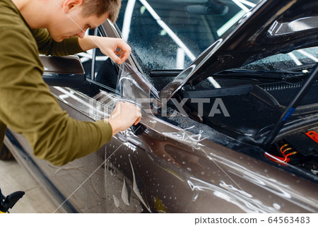 Worker installs protection film on car hood Worker installs protection film on car hood 64563483