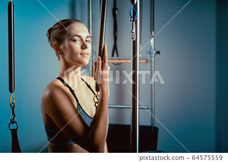 Young girl exercises on a bed of Pilates reformers, in the hall on a background of a gray wall. Fitness stretching concept. Healthy body, yoga, Pilates, copy space Young girl exercises on a bed of Pilates reformers, in the hall on a background of a gray wall. Fitness stretching concept. Healthy body, yoga, Pilates, copy space 64575559