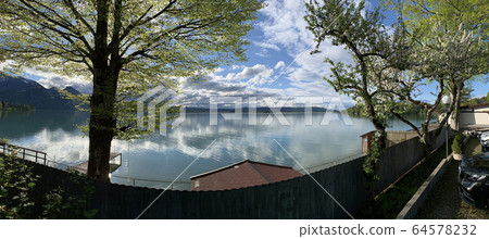 Coast one of lakes of the Italian Alps, quiet water, green trees. Reflection of clouds in water, a pier, mountains on a background 64578232