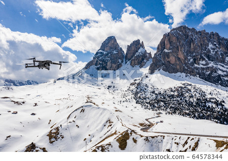 Italy, Dolomites, Canazei, The flying soaring drone hanged over mountains, on a background snow-covered mountains, the twisting road, the blue sky with white clouds, sunny weather, nobody Italy, Dolomites, Canazei, The flying soaring drone hanged over mountains, on a background snow-covered mountains, the twisting road, the blue sky with white clouds, sunny weather, nobody 64578344