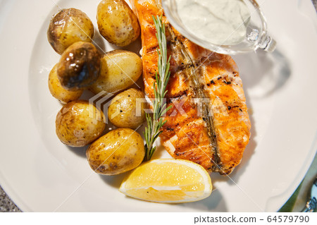 Grilled fish is on white plate with potatoes, lemon and sauce, Thyme branch, close up Grilled fish is on white plate with potatoes, lemon and sauce, Thyme branch, close up 64579790