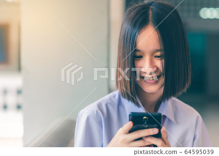 Smiling female Asian high school student in white uniform is enjoying social media on her smartphone. 64595039