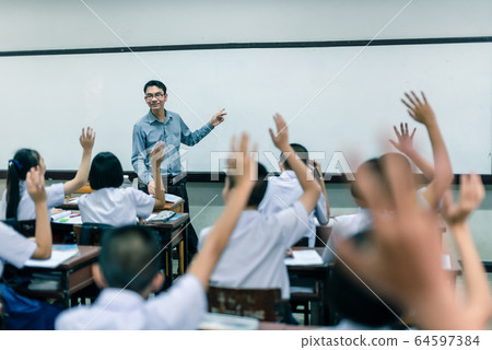 A smiling Asian male high school teacher teaches the white uniform students in the classroom by asking questions and then the students raise their hands for answers. A smiling Asian male high school teacher teaches the white uniform students in the classroom by asking questions and then the students raise their hands for answers. 64597384