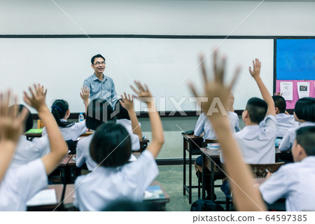 A smiling Asian male high school teacher teaches the white uniform students in the classroom by asking questions and then the students raise their hands for answers. A smiling Asian male high school teacher teaches the white uniform students in the classroom by asking questions and then the students raise their hands for answers. 64597385