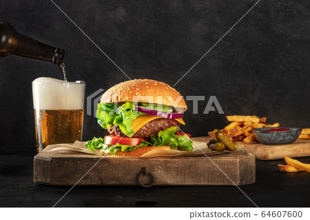 Burger and beer. Hamburger with beef, cheese, onion, tomato, and green salad, a side view on a dark background with copy space, gherkins, French fries and ketchup. Selective focus Burger and beer. Hamburger with beef, cheese, onion, tomato, and green salad, a side view on a dark background with copy space, gherkins, French fries and ketchup. Selective focus 64607600
