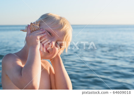 Cute blond boy put shell to his ear on sea 64609018