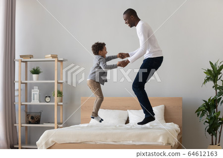 Happy black father and cute kid son jumping on bed 64612163