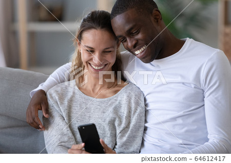 Happy mixed race couple using phone taking selfie on sofa 64612417