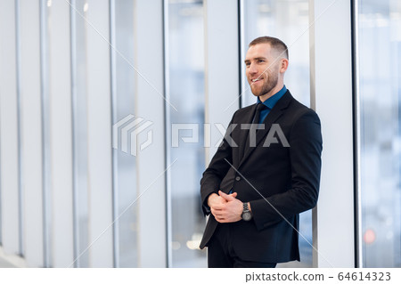 Stylish young businessman wearing a modern suit, who is a high achiever, standing on the top floor of an office building looking out at the view through large windows Stylish young businessman wearing a modern suit, who is a high achiever, standing on the top floor of an office building looking out at the view through large windows 64614323