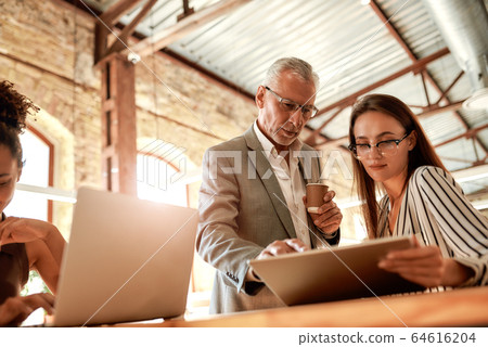 Sharing experience. Senior man in formal wear holding coffee cup and explaining something to his young female colleague while working together in the modern office Sharing experience. Senior man in formal wear holding coffee cup and explaining something to his young female colleague while working together in the modern office 64616204