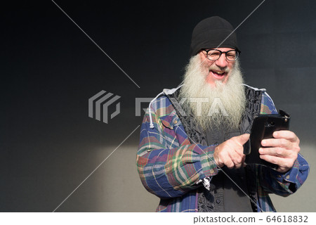 Happy mature bearded hipster man using phone against black wall 64618832