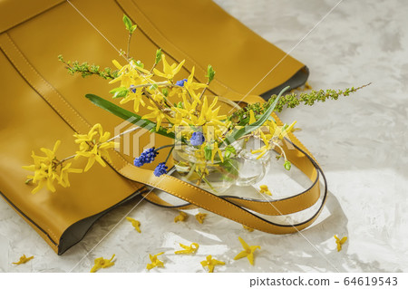 Lying on a light background yellow handbag and bouquet of blue and yellow flowers in vase close-up, top view, selective focus. Concept of spring or summer shopping 64619543