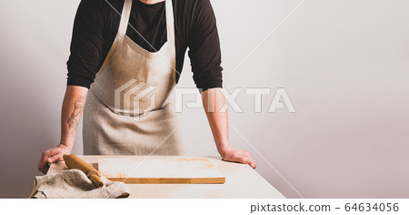 Men cook in a cotton apron holds a wooden rolling pin at the kitchen table with a cutting board. Rustic style. 64634056