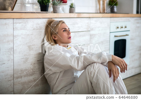 Blonde woman in white workwear sitting on the floor and feeling unwell 64646880