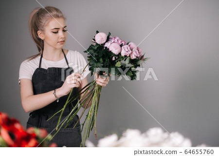 Lovely young woman florist wearing apron making floral composition from fresh roses Lovely young woman florist wearing apron making floral composition from fresh roses 64655760