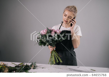 Busy young woman florist wearing apron talking on mobile phone with client Busy young woman florist wearing apron talking on mobile phone with client 64655768
