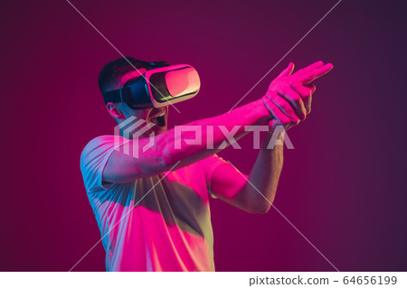 Caucasian man's portrait isolated on pink-purple studio background in neon light, playing with VR-headset 64656199
