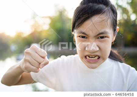 Serious asian child girl with her fist raised for threat warning or quarreling,angry female threatening with fist,about to punch,aggressive schoolgirl,facial expression,concept of aggression,bully  64656458