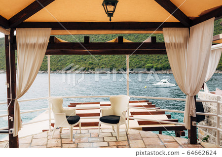 Two white chairs with black pillows stand in a gazebo by the sea overlooking a white boat, in Montenegro, on the Lustica Peninsula Two white chairs with black pillows stand in a gazebo by the sea overlooking a white boat, in Montenegro, on the Lustica Peninsula 64662624