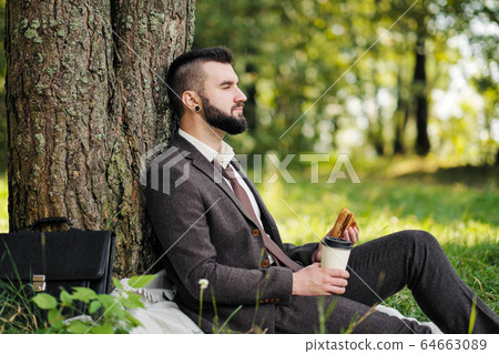 Young attractive bearded business man sitting on green grass under tree and resting in park. Drink coffee and eat sandwich. Relaxed, tired of work, lunch break. Young attractive bearded business man sitting on green grass under tree and resting in park. Drink coffee and eat sandwich. Relaxed, tired of work, lunch break. 64663089