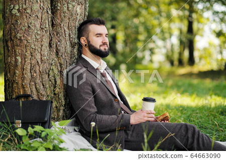 Young attractive bearded business man sitting on green grass under tree and resting in park. Drink coffee and eat sandwich. Relaxed, tired of work, lunch break. 64663090