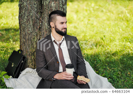 Young attractive bearded business man sitting on green grass under tree and resting in park. Drink coffee and eat sandwich. Relaxed, tired of work, lunch break. 64663104