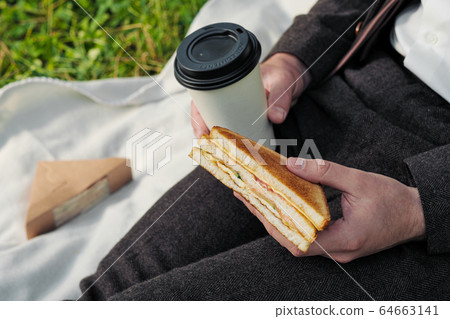 Hands of a man in a suit with a glass of takeaway coffee and a triangular sandwich with tuna. 64663141