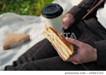 Hands of a man in a suit with a glass of takeaway coffee and a triangular sandwich with tuna. 64663142