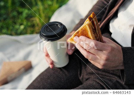 Hands of a man in a suit with a glass of takeaway coffee and a triangular sandwich with tuna. 64663143