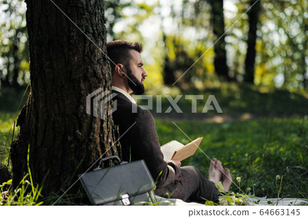 Young attractive bearded business man sitting on green grass under tree and resting in park. Read book, drink coffee. Relaxed, tired of work, lunch break. 64663145
