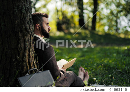 Young attractive bearded business man sitting on green grass under tree and resting in park. Read book, drink coffee. Relaxed, tired of work, lunch break. 64663146