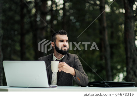 Young attractive business man in suit and tie sits at desk and works on computer outdoors. Drink coffee from white cup. Green trees, nature and park on background. Young attractive business man in suit and tie sits at desk and works on computer outdoors. Drink coffee from white cup. Green trees, nature and park on background. 64678319