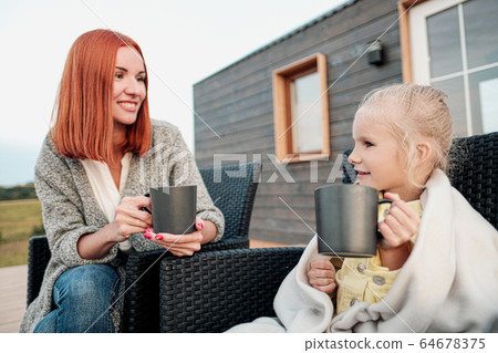 A young mother and little daughter sit in chairs in warm rugs and drink tea from the mugs on the terrace of a wooden house. Autumn evening in the country. 64678375