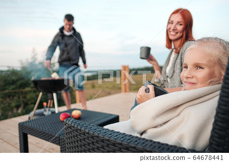 A young Caucasian family couple with a baby daughter sits on the terrace in chairs and drinks hot drinks. Against the background of a small modern rustic wooden house. Father is cooking barbecue. 64678441