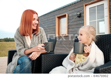 A young mother and little daughter sit in chairs in warm rugs and drink tea from the mugs on the terrace of a wooden house. Autumn evening in the country. A young mother and little daughter sit in chairs in warm rugs and drink tea from the mugs on the terrace of a wooden house. Autumn evening in the country. 64678902