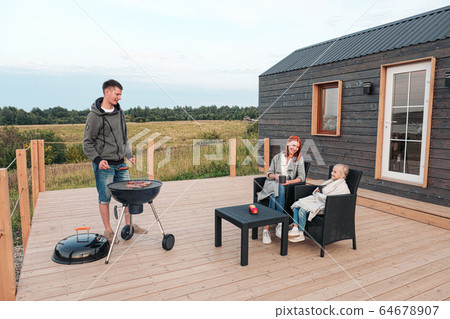 A young Caucasian family couple with a baby daughter sits on the terrace in chairs and drinks hot drinks. Against the background of a small modern rustic wooden house. Father is cooking barbecue. 64678907