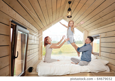 Young Caucasian family couple with baby daughter in a small modern rustic house with a large window. Lying on the bed, hugging, playing and looking out the window. 64679032